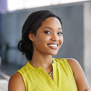 The image shows a woman with dark hair, wearing a yellow top and a necklace, smiling at the camera.